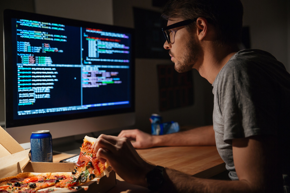 Profile of concentrated young software developer eating pizza and coding at home Profile of concentrated young software developer eating pizza and coding at home